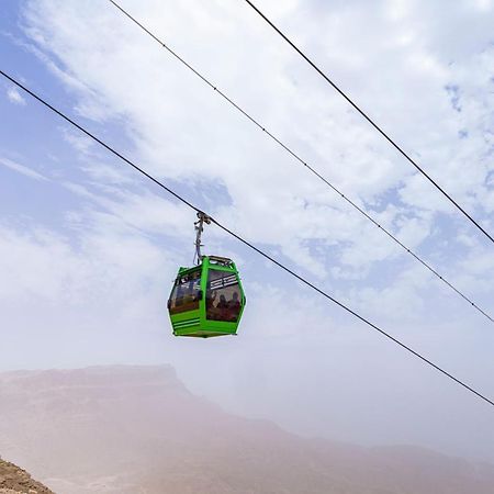 加拉拉马加斯迪克托利普度假村 艾因苏赫纳 外观 照片 The photo shows a green cable car suspended in the air, traveling along overhead cables. The backdrop features a hazy landscape, likely mountainous or elevated terrain, with a cloudy sky. The scene suggests a scenic transportation method, possibly us